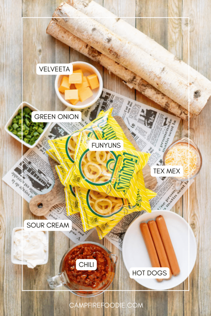 Top-down view of a wooden table with labeled ingredients: Velveeta cubes, green onions, Funyuns chips, shredded Tex Mex cheese, sour cream, chili, and hot dogs, along with birch logs and newspaper.