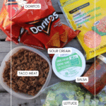 Overhead view of taco salad ingredients: bags of Doritos chips, shredded cheddar cheese, cooked taco meat in a container, sour cream, salsa in a small container, and chopped lettuce on a wooden surface.
