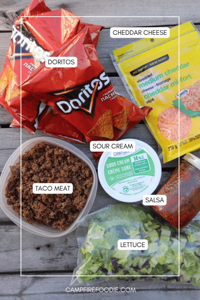 Overhead view of taco salad ingredients: bags of Doritos chips, shredded cheddar cheese, cooked taco meat in a container, sour cream, salsa in a small container, and chopped lettuce on a wooden surface.