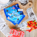 Overhead view of whipped cream, mini Oreo packs on a wooden board, chocolate pudding cups, and gummy worms, all labeled, on a rustic wood table with newspaper sheets underneath.