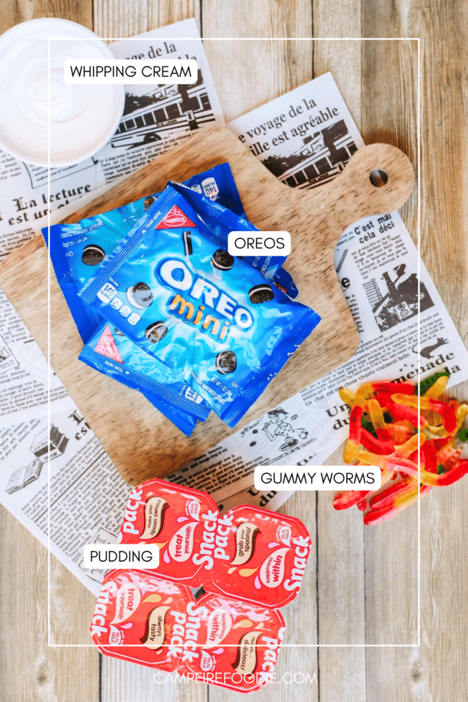 Overhead view of whipped cream, mini Oreo packs on a wooden board, chocolate pudding cups, and gummy worms, all labeled, on a rustic wood table with newspaper sheets underneath.