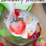 Top view of a plastic pouch filled with strawberry shortcake dessert, including strawberries, whipped cream, cake pieces, and a spoon. Sliced strawberries and a dessert package are visible on a wooden surface nearby. Text overlays read: Walking Dessert strawberry shortcake and campfirefoodie.com.