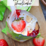 Open pudding cup topped with whipped cream, strawberry sauce, a sliced strawberry, and crumbled biscuits, with a plastic spoon inside. Fresh strawberries are nearby on a wooden surface. Text reads: WALKING DESSERT strawberry shortcake.