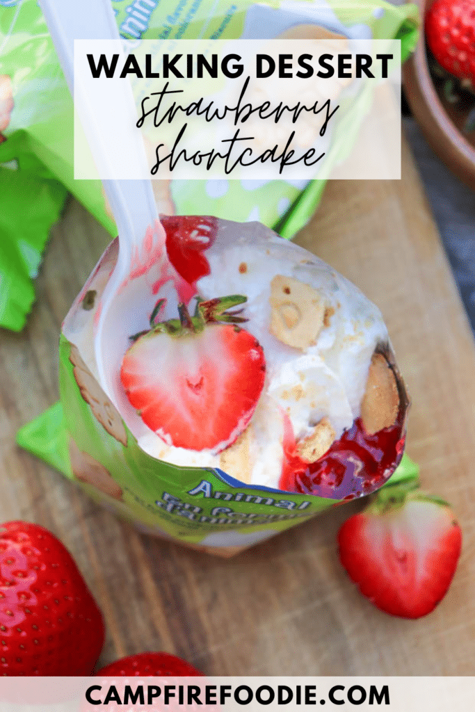 Open pudding cup topped with whipped cream, strawberry sauce, a sliced strawberry, and crumbled biscuits, with a plastic spoon inside. Fresh strawberries are nearby on a wooden surface. Text reads: WALKING DESSERT strawberry shortcake.