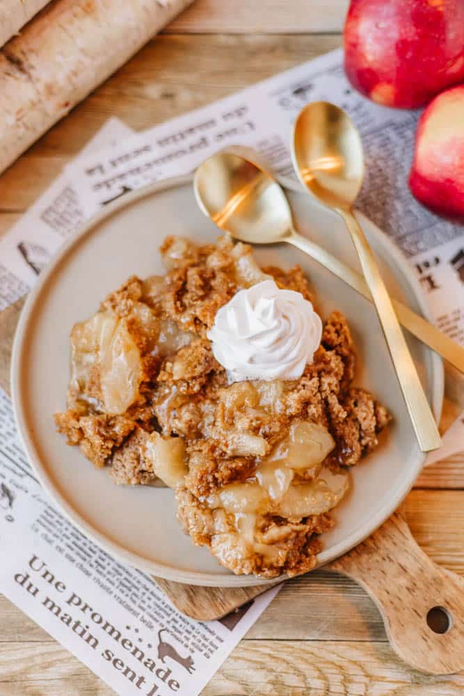 A plate of apple crumble topped with whipped cream is served on a wooden board. Two gold spoons are placed beside it, reminiscent of a Dutch oven peach cobbler. Red apples and a rolling pin are in the background, along with newspaper pages.