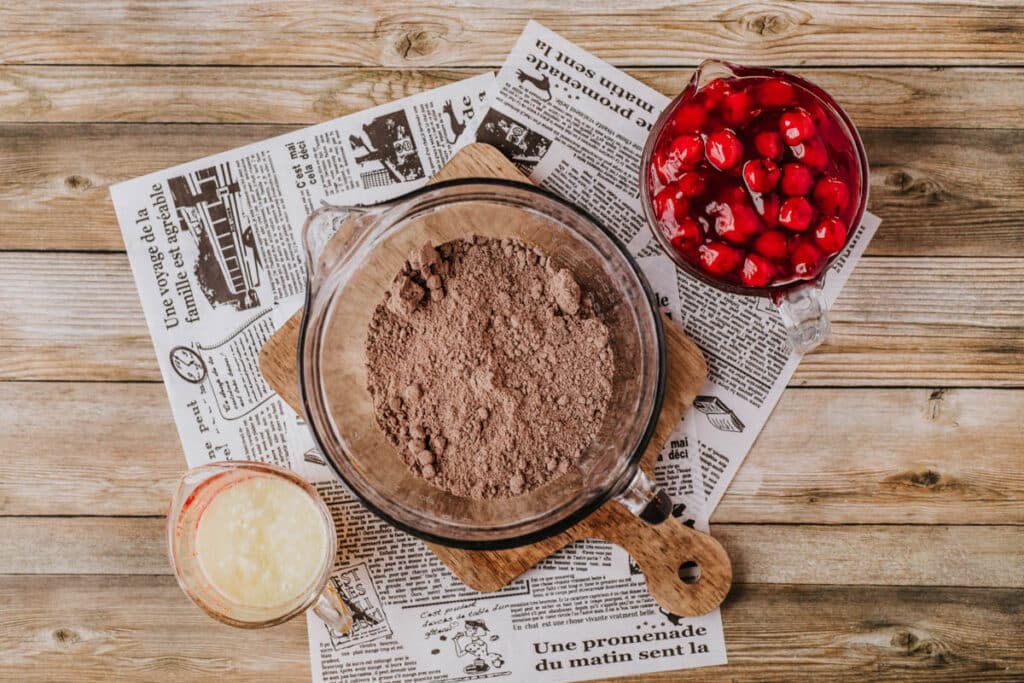 A wooden table is graced with a glass bowl of cocoa powder, a pitcher of red cherries, and a glass measuring cup of wet ingredients, all artfully arranged on sheets of newspaper, as if waiting to join the delightful aroma of a Dutch oven peach cobbler.