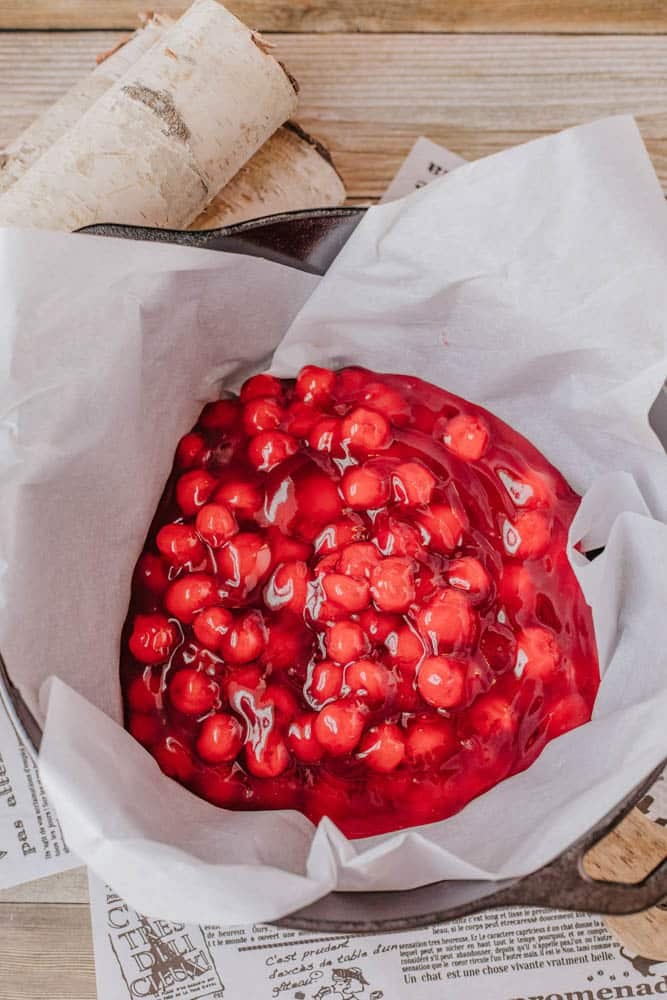 Cherries in a vibrant red sauce sit atop parchment paper in a seasoned cast iron skillet. The skillet, reminiscent of preparing a classic Dutch oven peach cobbler, rests on sheets of newspaper with a wooden surface behind. A piece of birch bark is partially visible at the top.