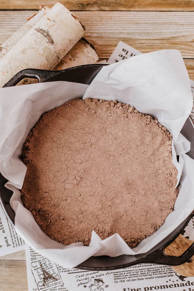A black baking dish lined with parchment paper is filled with a crumbly chocolate pastry dough, reminiscent of the rustic charm found in a Dutch oven peach cobbler. It sits on sheets of printed newspaper, while pieces of birch bark add a natural touch to the wooden surface.