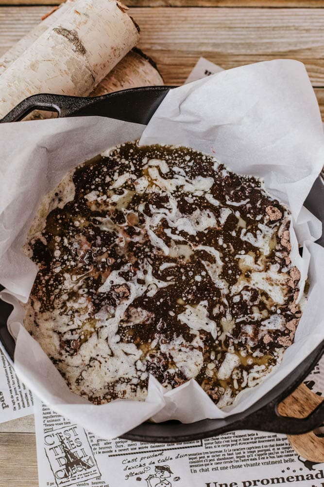 A round chocolate dessert with a marbled appearance sits elegantly in a parchment-lined black pan, reminiscent of the warmth youd find in a dutch oven peach cobbler. Its placed on a rustic wooden table with newspaper pages beneath, beside a birch bark log.
