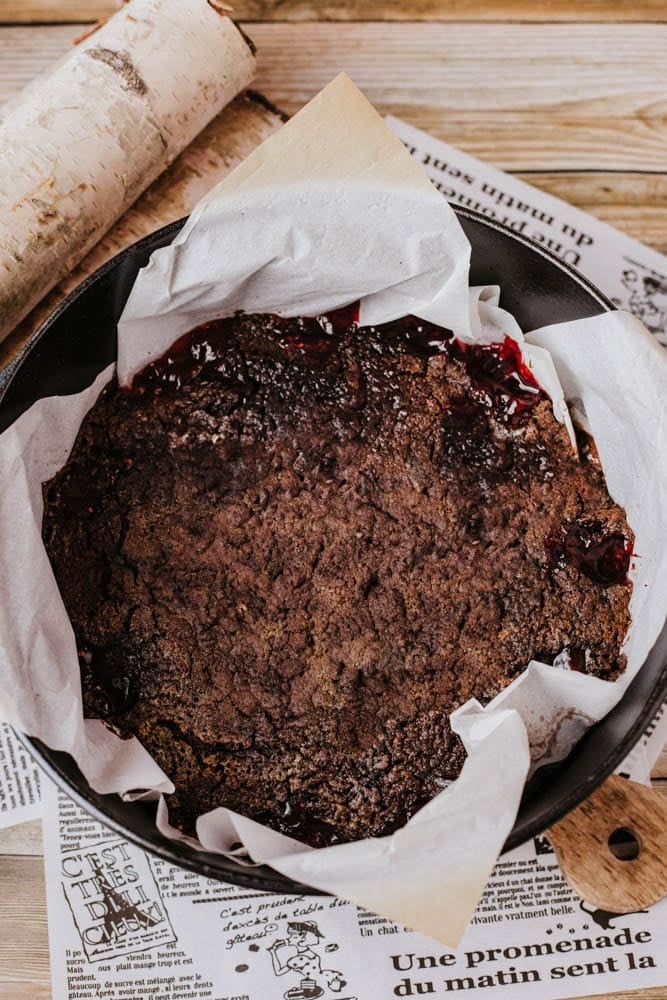 A round, rustic berry crumble in a black cast iron pan lined with parchment paper echoes the charm of a traditional Dutch oven peach cobbler. The dark baked topping is slightly cracked, revealing red berry fillings, as it rests on newspapers against a wooden table backdrop.