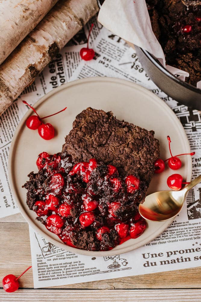 A plate with chocolate crumble dessert topped with bright red cherries, placed on newspaper. A gold spoon rests on the plate. In the background, theres a dish reminiscent of a Dutch oven peach cobbler along with birch logs for a rustic touch.