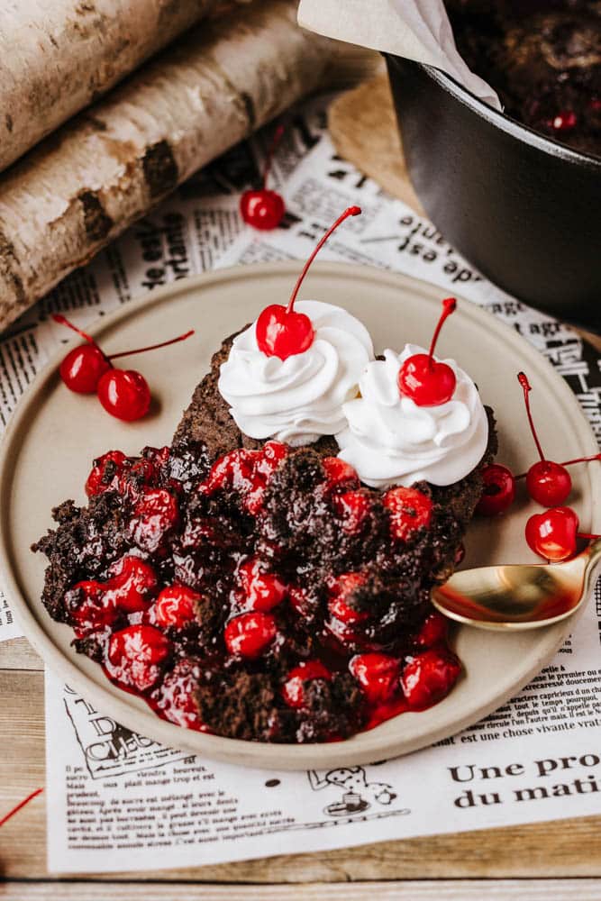 A plate of chocolate cake topped with cherry pie filling, whipped cream, and maraschino cherries rests on a table covered with newspapers. Beside it lies a gold spoon, while in the background, partially visible logs and a dutch oven hint at peach cobbler delights yet to come.