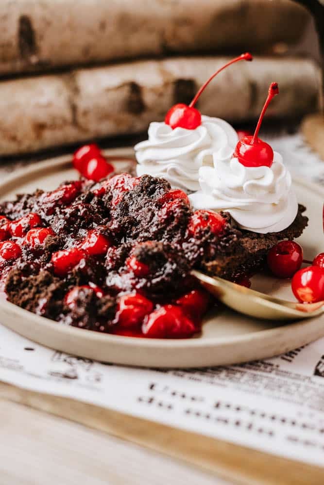 A close-up of a chocolate dessert topped with cherries and whipped cream on a plate, reminiscent of the comfort found in a warm Dutch oven peach cobbler. Each dollop of whipped cream cradles a cherry, surrounded by more cherries. A gold spoon rests nearby, while the background is blurred.