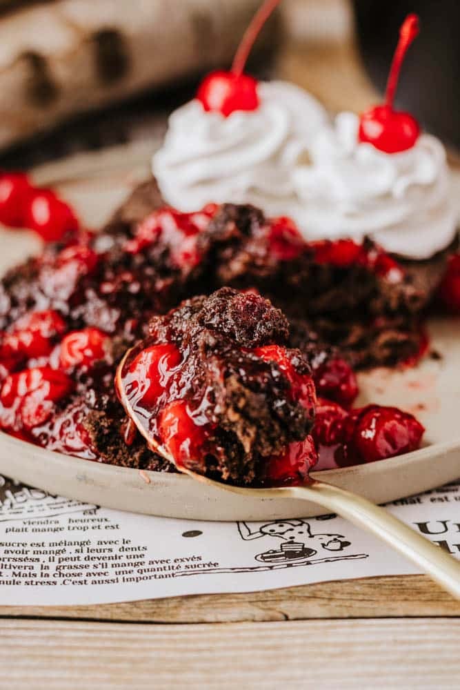 A plate with rich chocolate dessert topped with vibrant red cherries and whipped cream sits beside a luscious dutch oven peach cobbler. A spoonful of the chocolate delight, showing gooey cherry filling, is placed next to it. Both rest on a newspaper page, adding a rustic touch.