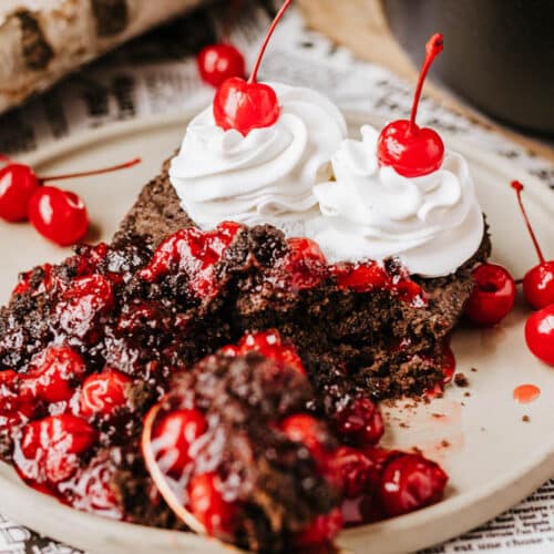 A close-up of a chocolate cake topped with whipped cream and cherries, surrounded by scattered cherries. A piece is cut out, revealing its rich texture on a plate with a vintage newspaper beneath. Logs and the aroma of dutch oven peach cobbler linger in the background where a black pot rests.