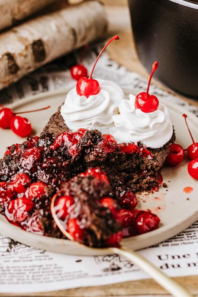 A close-up of a chocolate cake topped with whipped cream and cherries, surrounded by scattered cherries. A piece is cut out, revealing its rich texture on a plate with a vintage newspaper beneath. Logs and the aroma of dutch oven peach cobbler linger in the background where a black pot rests.