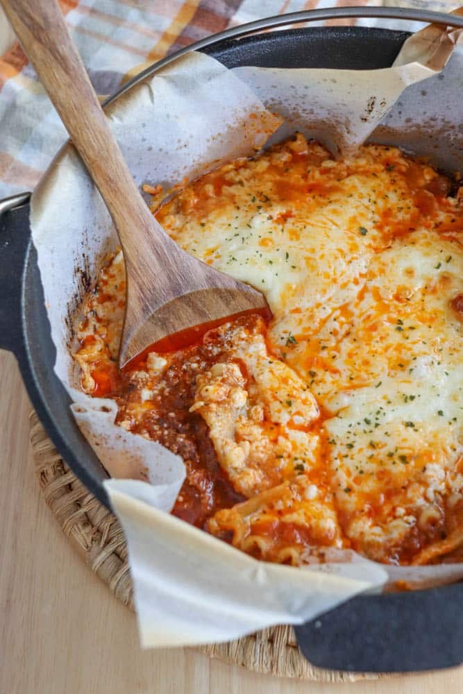 A wooden spoon scoops a cheesy, baked dutch oven lasagna recipe from a parchment-lined cast iron pot, with gooey cheese and tomato sauce visible. The dish rests on a woven mat, with a plaid cloth in the background.