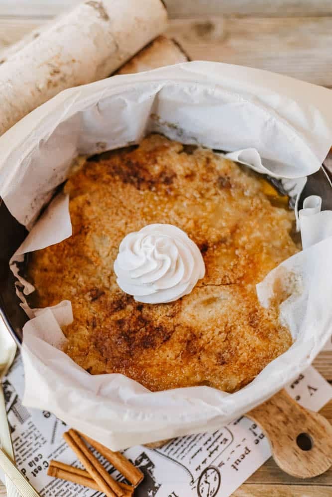 A baked dessert in a round dish lined with parchment paper, this Dutch oven peach cobbler is topped with a dollop of whipped cream. The surface is golden brown and crusted. Cinnamon sticks and rolled parchment lie nearby on a wooden table.