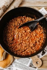 A pot of Dutch oven sloppy joes with ground beef in tomato sauce is being stirred with a black spoon, resting on a wooden table beside hamburger buns and newspaper.