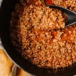 A close-up of seasoned ground beef in tomato sauce cooking in a black dutch oven, with a black spoon scooping some up for homemade sloppy joes. Hamburger buns are partially visible on the side, sitting on newspaper.
