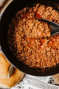 A close-up of seasoned ground beef in tomato sauce cooking in a black dutch oven, with a black spoon scooping some up for homemade sloppy joes. Hamburger buns are partially visible on the side, sitting on newspaper.
