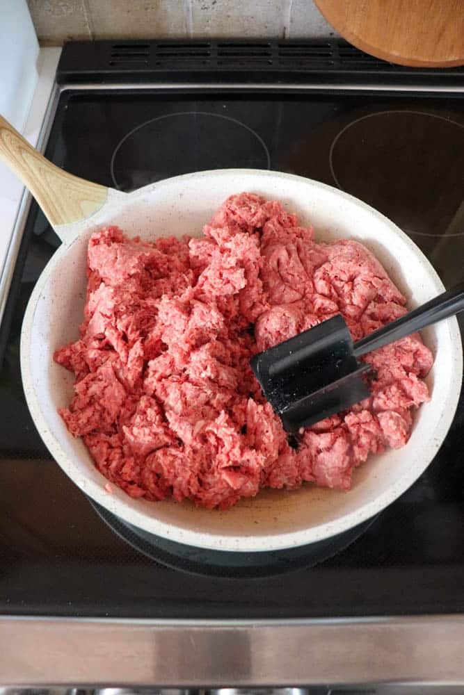 A white skillet containing raw ground beef is placed on a stovetop, with a black spatula and a wooden-handled utensil resting in the pan, ready to cook the meat.