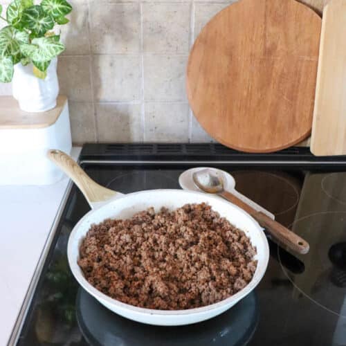 A white skillet filled with cooked ground beef sits on a stovetop, with a wooden spoon resting on the pan. Cutting boards and a potted plant are in the background on a kitchen counter.