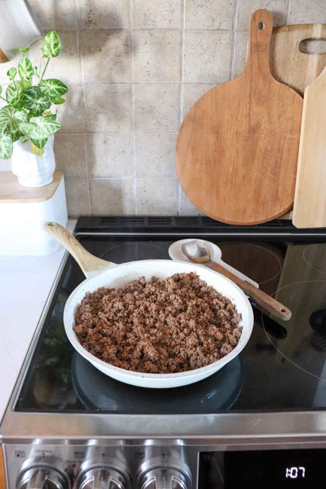 A white skillet filled with cooked ground beef sits on a stovetop, with a wooden spoon resting on the pan. Cutting boards and a potted plant are in the background on a kitchen counter.