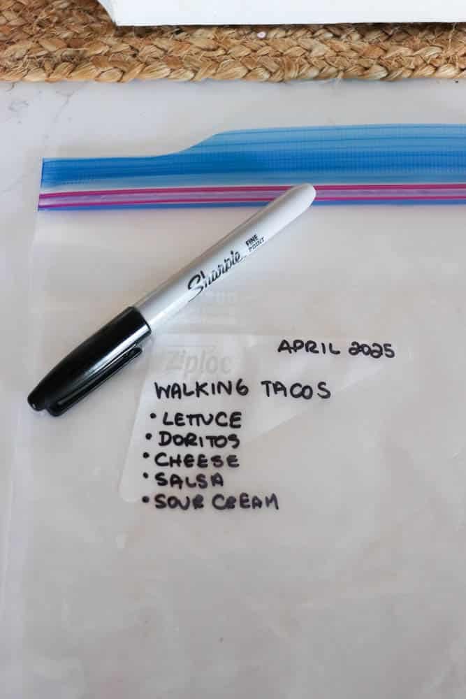 A Sharpie marker rests on a Ziploc bag labeled April 2025, Walking Tacos with a list: lettuce, Doritos, cheese, salsa, sour cream. The bag is on a white surface with a woven mat partially visible.
