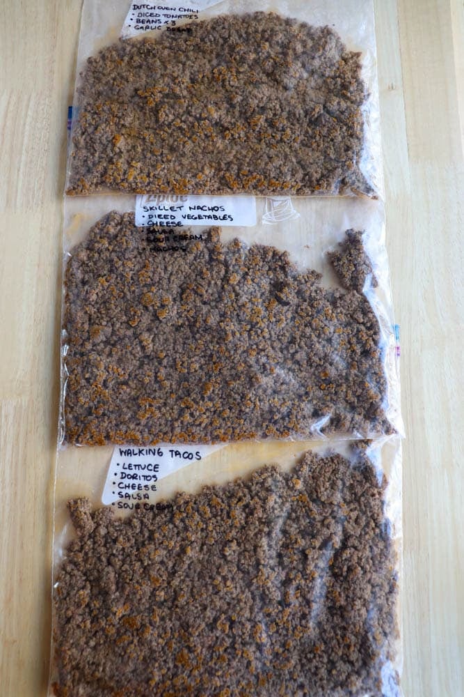 Three plastic bags filled with cooked ground beef are laid out on a wooden surface. Each bag is labeled with different meal options written in marker, such as Dutch oven chili and walking tacos.