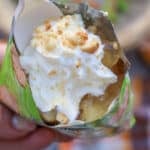A close-up of a hand holding a bag filled with a dessert topped with whipped cream and crushed cookies, with a plastic spoon inserted. The bag is green with colorful designs.
