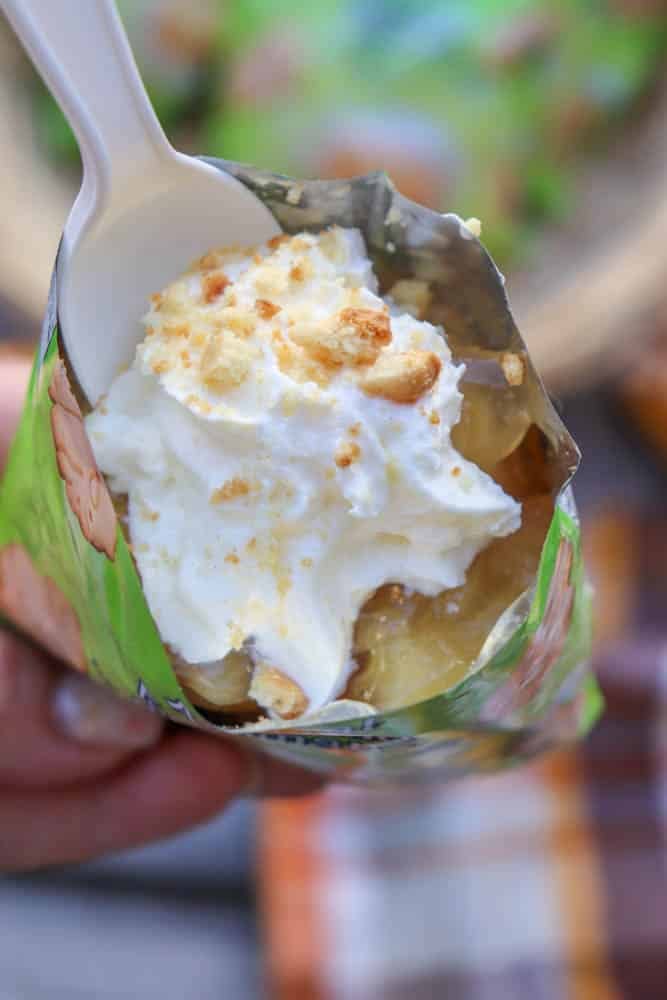 A close-up of a hand holding a bag filled with a dessert topped with whipped cream and crushed cookies, with a plastic spoon inserted. The bag is green with colorful designs.
