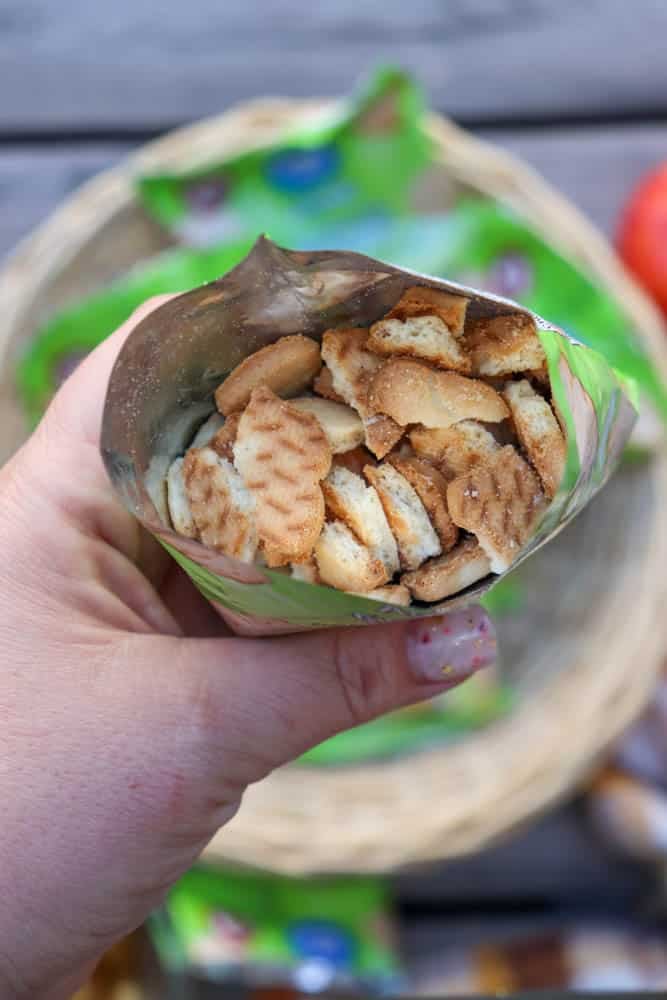 A hand holding an open green snack bag filled with small, round, golden cookies above a wicker basket.