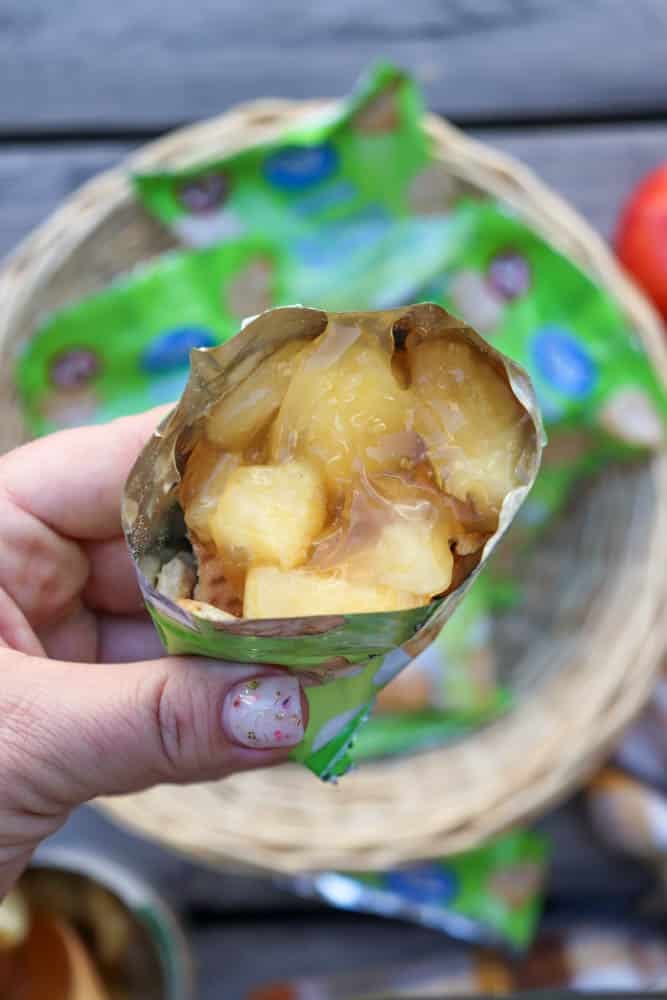 A hand holding an open snack pouch filled with chunks of pineapple and juice. A woven basket and green packaging are blurred in the background.