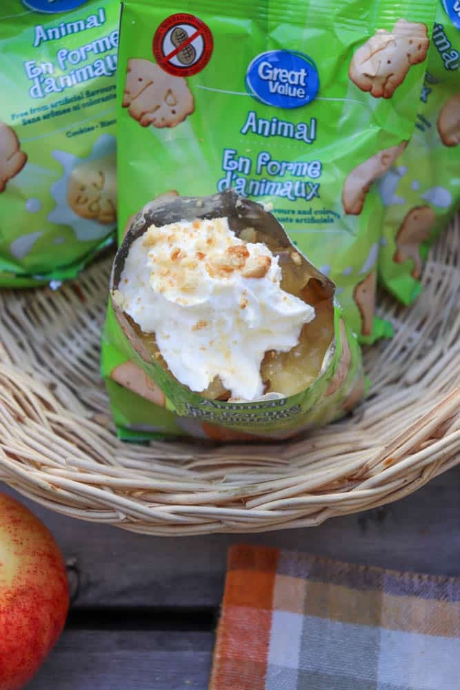 A small bag of animal-shaped cookies is used as a container for apple pie filling and whipped cream, sitting in a woven basket with other unopened cookie bags nearby. An apple and a plaid cloth are also visible.
