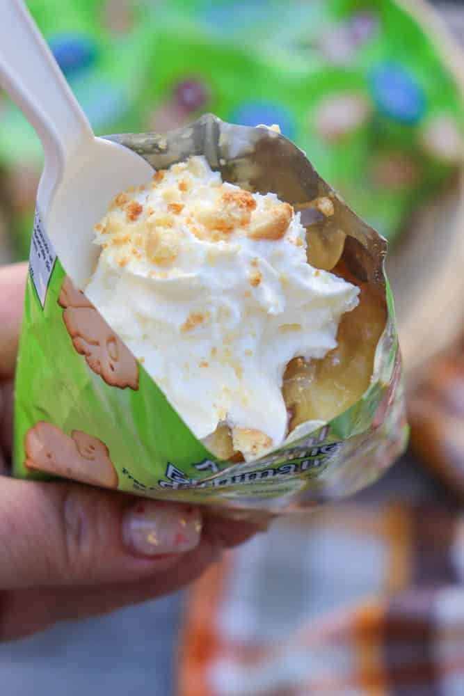 A hand holds an opened green snack bag filled with banana pudding, whipped cream, and crumbled cookies. A white plastic spoon is inserted into the dessert.