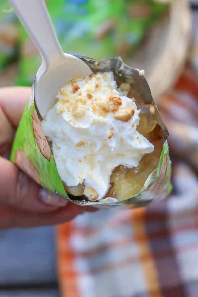 A hand holds an opened snack-sized bag filled with banana pudding, topped with whipped cream and crumbled cookies, with a white plastic spoon inserted. A plaid cloth is blurred in the background.