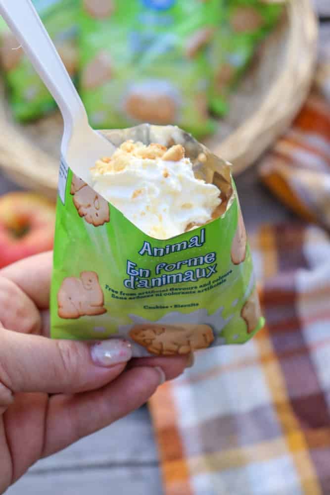 A hand holds an open green bag of animal crackers, topped with whipped cream and crumbled cookies, with a white plastic spoon inside. A checkered cloth and snacks are blurred in the background.