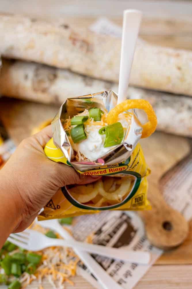 A hand holds an open bag of onion flavored rings topped with shredded cheese, sour cream, and chopped green onions, with a white plastic fork inside.
