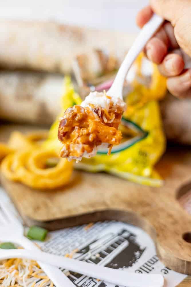 A hand holds a white plastic fork with a bite of cheesy, loaded Fritos topped with ground meat, sour cream, and shredded cheese. A yellow Fritos bag, cutting board, and onion rings are visible in the background.