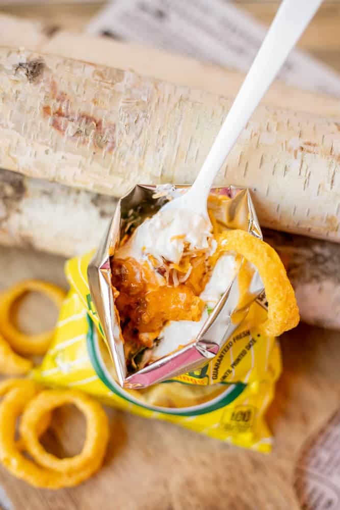 A small open bag of potato chips topped with sour cream, shredded cheese, and an onion ring, with a white plastic fork sticking out. Several onion rings are scattered nearby on a wooden surface.