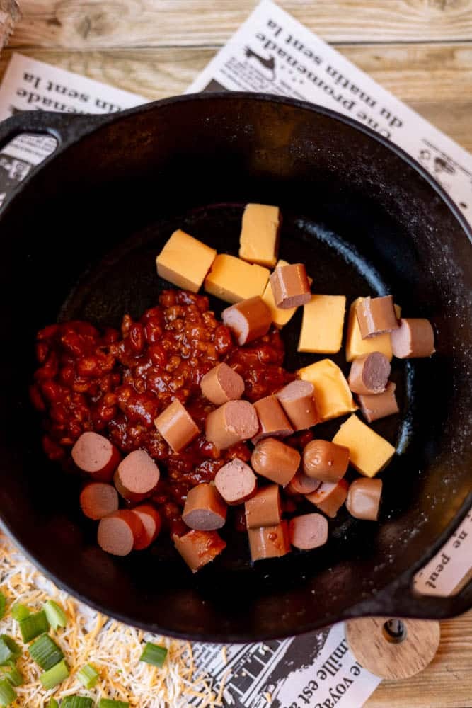 A black cast iron skillet contains baked beans, cubed yellow cheese, and sliced hot dogs, ready to be cooked. The skillet sits on a newspaper-lined wooden surface with chopped green onions and shredded cheese nearby.