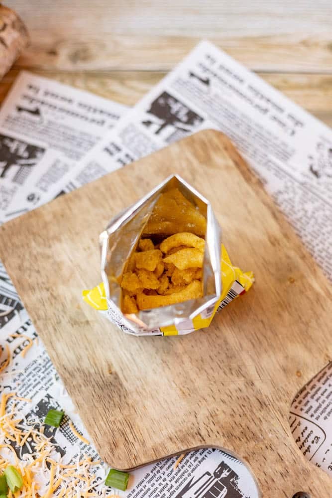 An open bag of corn chips sits on a wooden cutting board, surrounded by shredded cheese, green onions, and newspaper sheets on a rustic table.