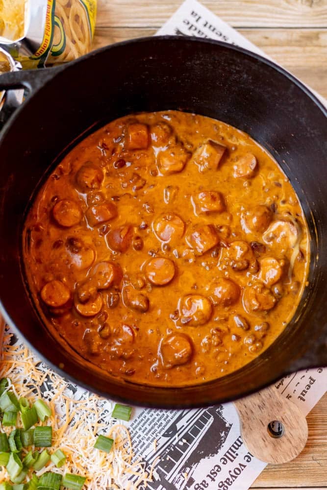 A pot of creamy, chunky sausage and bean stew sits on a wooden table, with shredded cheese and chopped green onions nearby as toppings. The stew has a rich, orange-brown color and visible sausage slices.
