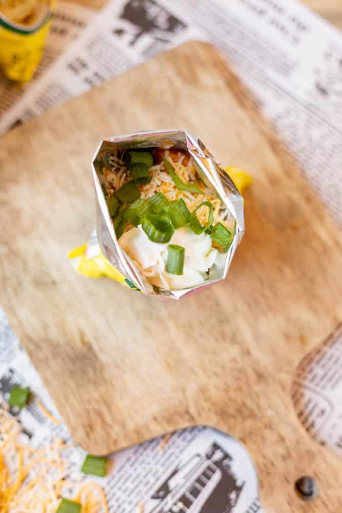 An open snack-size chip bag filled with shredded cheese, chopped green onions, and other toppings sits on a wooden board over newspaper. Some cheese and green onion pieces are scattered nearby.