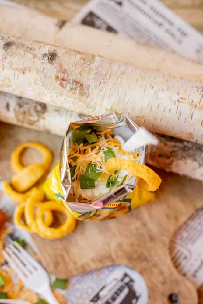 An open snack-size chip bag filled with shredded cheese, lettuce, and diced tomatoes, with a plastic fork sticking out. Curly chips and utensils are scattered nearby on a rustic wooden surface.
