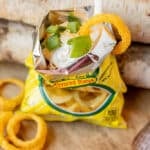 A small yellow bag of onion-flavored rings is opened and topped with sour cream and chopped green onions, with a plastic spoon and an onion ring resting on the edge of the bag.