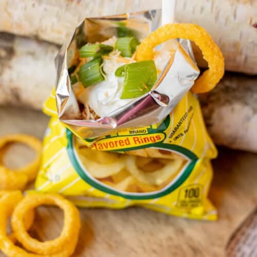 A small yellow bag of onion-flavored rings is opened and topped with sour cream and chopped green onions, with a plastic spoon and an onion ring resting on the edge of the bag.
