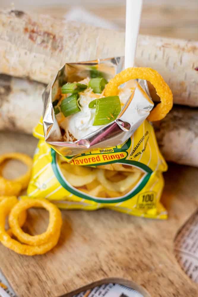 A small yellow bag of onion-flavored rings is opened and topped with sour cream and chopped green onions, with a plastic spoon and an onion ring resting on the edge of the bag.