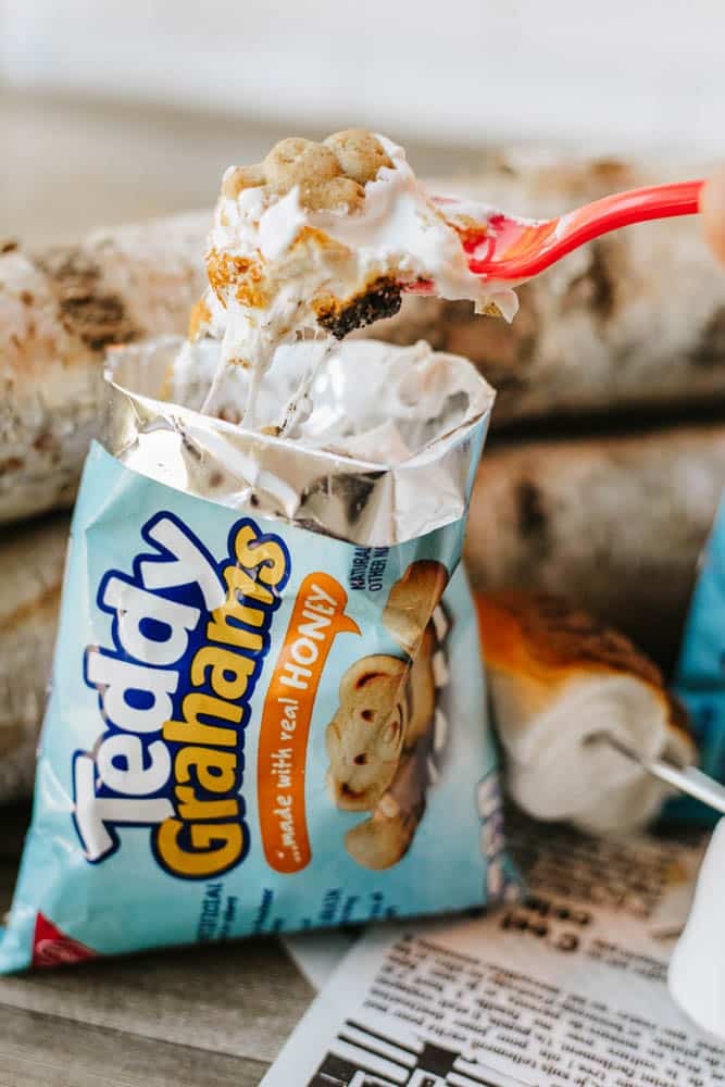 A red spoon scoops marshmallow and chocolate from an open Teddy Grahams honey snack bag, creating a makeshift s’mores treat; logs and packaging are visible in the background.
